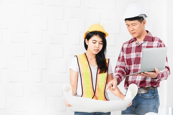 Middle aged Asian architect, in jeans and checked shirt, showing data on laptop to young female Asian assistant, wearing orange, yellow reflective safety vest, and hardhat, blueprints in hands