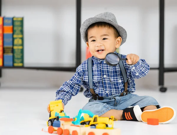 Bir yaşındaki Asyalı erkek bebek gülümseme ve büyüteç için tutarak katta oturan oyuncaklar mutlu kitap odasında zevk oynamak.