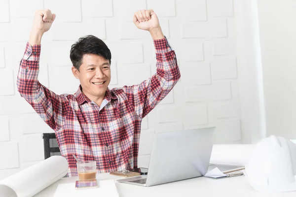 Yumruk kadar dizüstü bilgisayar, kask, tablet, kahve ve belgeleri arka planda tutan kazanan jest yapma inşaat site ofiste çalışan Asya yakışıklı mimar, ekose desenli gömlek, orta yaşlı