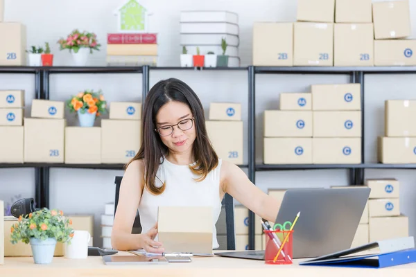 Portrait of young, beautiful, Asian business lady, long black hair, wearing eyeglasses, in casual fashion, check delivery tracking reference number on notebook, with startup home office background
