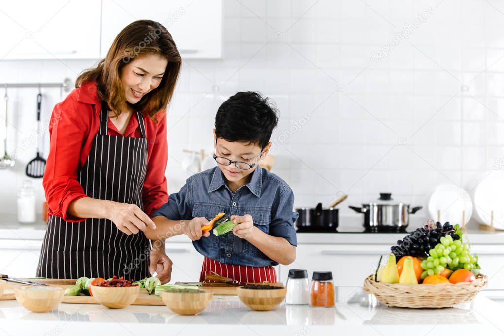 Asian family mom in red shirt and black  apron is teaching her son use  tool to peel fruit and vegetables to prepare food in white clean modern kitchen.