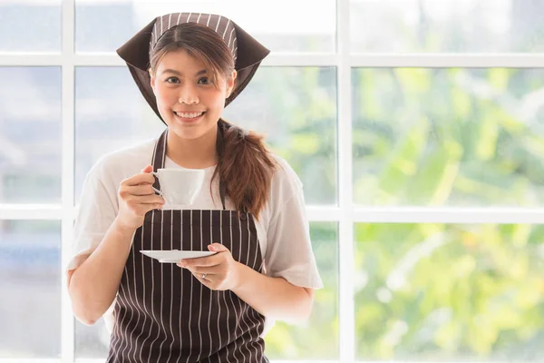縞模様の料理の帽子とエプロンで幸せな若いと美しいアジアの女性のフロントビューの肖像画 大きなガラス窓でモダンなキッチンの背景でコーヒーブレイクをお楽しみください カメラで笑顔 — ストック写真