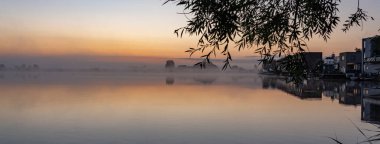 Güneşin doğuşundan hemen önce, rüzgarsız ve sisli bir sabahta Noordhovense plakalarındaki evler suya yansıyor.