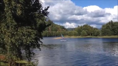 Gökyüzünde bulutlar olan bir nehir manzarası, çok uzakbir yelkenlide bir kız ve ön planda büyük bir ağaç.