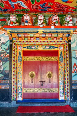 Renkli dekore edilmiş, Rumtek Budist manastırının (Sikkim, Hindistan) bir tapınağının mabedine ustaca yapılmış ve süslü kapı tokmakları ile, boyalı duvarlar ve tavan, kapının üzerinde beyaz aslan heykelleri.