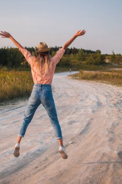 Gül gömlekli, kot pantolonlu ve bej renkli kovboy şapkalı bir kadın gün batımında bir tarlada duruyor. Kız doğal olarak eğleniyor.
