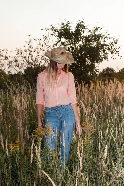 Woman in a rose shirt, jeans and a beige cowboy hat is standing in a field on a sunset time. The girl is having fun in a nature