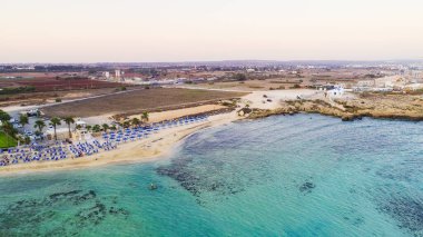 Kıyı şeridi günbatımı ve tarihi plaj manzarası yukarıdan Agia Thekla, Ayia Napa, Famagusta, Kıbrıs. Kuş bakışı gökyüzü manzarası turistlerin ilgi odağı altın kumlu körfez, adacık, Ammochostos 'taki güneşli yataklar..