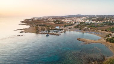 Kıbrıs Rum Kesimi 'nin Protaras, Famagusta, Agia Triada plajındaki kıyı şeridi günbatımı ve tarihi beyaz şapel manzarası. Gündoğumunda Ammochostos semtinde Ayia Trias Körfezi kilisesi, kum yatakları ve güneş yatakları kuşbakışı turizm manzarası.