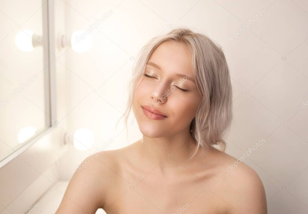 Beautiful young woman with clean fresh skin and eyes closed sitting in white studio with mirror