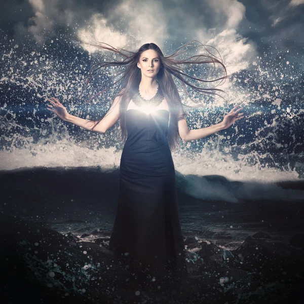 beautiful woman with long flying hair and glowing light in her heart standing with spread hands on dark stormy ocean and dramatic sky background