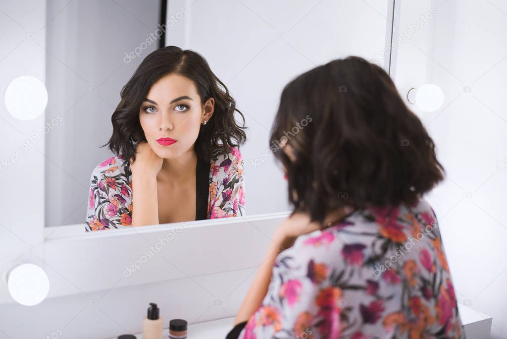 Young attractive brunette model sits in front of mirror in a bathrobe and looks at her reflection in a white dressing room surrounded by bright bulbs. Body positive concept, confirmation of its beauty