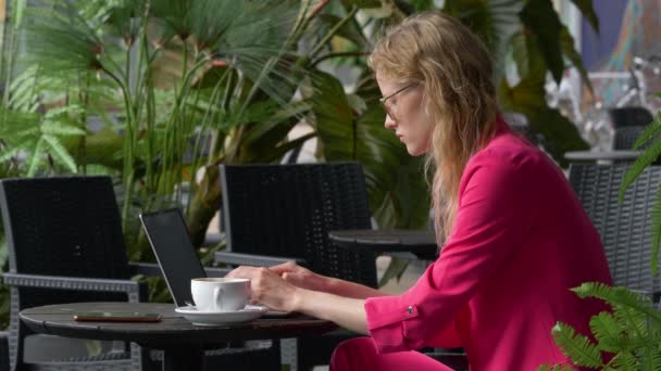 La mujer adicta al trabajo bebe su café de la mañana y termina sus sábanas para la reunión de la mañana. Mujer de negocios con traje rosa rodeada de plantas verdes. Rubia pelo ondulado, gafas, pecas . — Vídeos de Stock