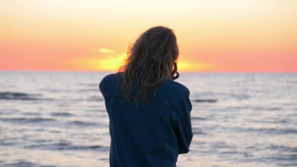 Närbild skott av kvinna stående och titta på solnedgången nära havet. Vackert blont vågigt hår skakar i vinden. Ur ett perspektiv. Avslappnad kvinna njuter av sin frihet. — Stockvideo