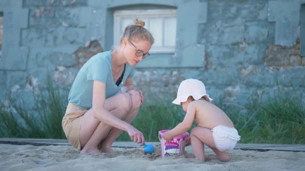 家族の時間だ。若い幸せな母親は砂浜で砂と彼女の娘と遊ぶ。幼児の女の子は太陽から白い帽子を身に着けている。母は砂の城を構築する方法を示しています. — ストック動画