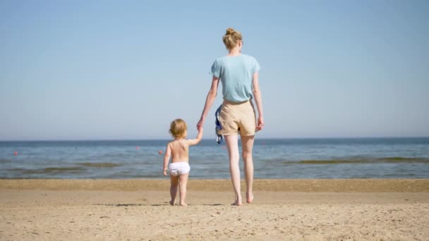 Macierzyństwo. Młoda matka idzie ze swoją córeczką nad morze. Blondynka trzyma córkę za rękę. Powoli spacerując po plaży. — Wideo stockowe