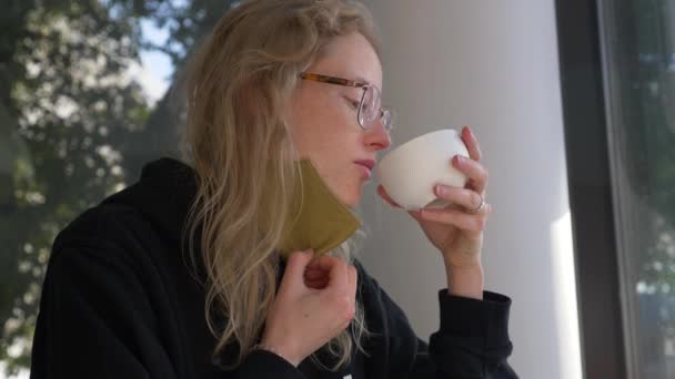 Una joven rubia bebe café durante la cuarentena. Mujer somnolienta con mascarilla intenta disfrutar de su rutina matutina. — Vídeos de Stock