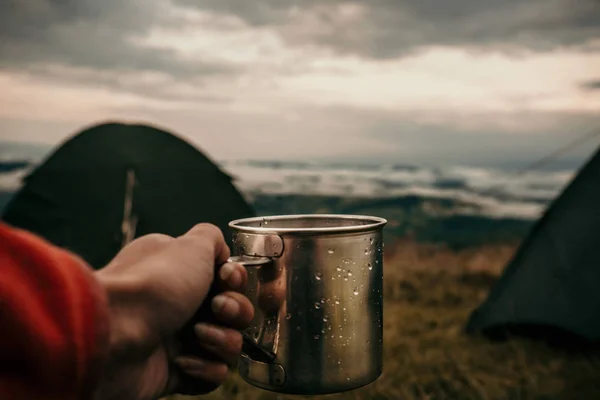 Kubek metalowy z herbatą w ręku turysty w obozie namiotowym w górach — Zdjęcie stockowe