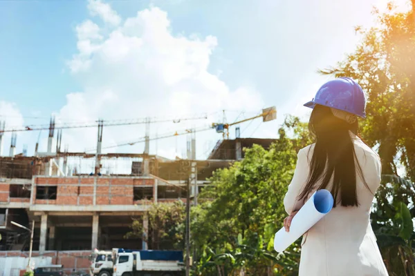 Asian woman architect wearing blue safety helmet checking working progress at contruction site.