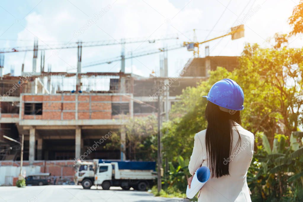 Asian woman architect wearing blue safety helmet checking working progress at contruction site.