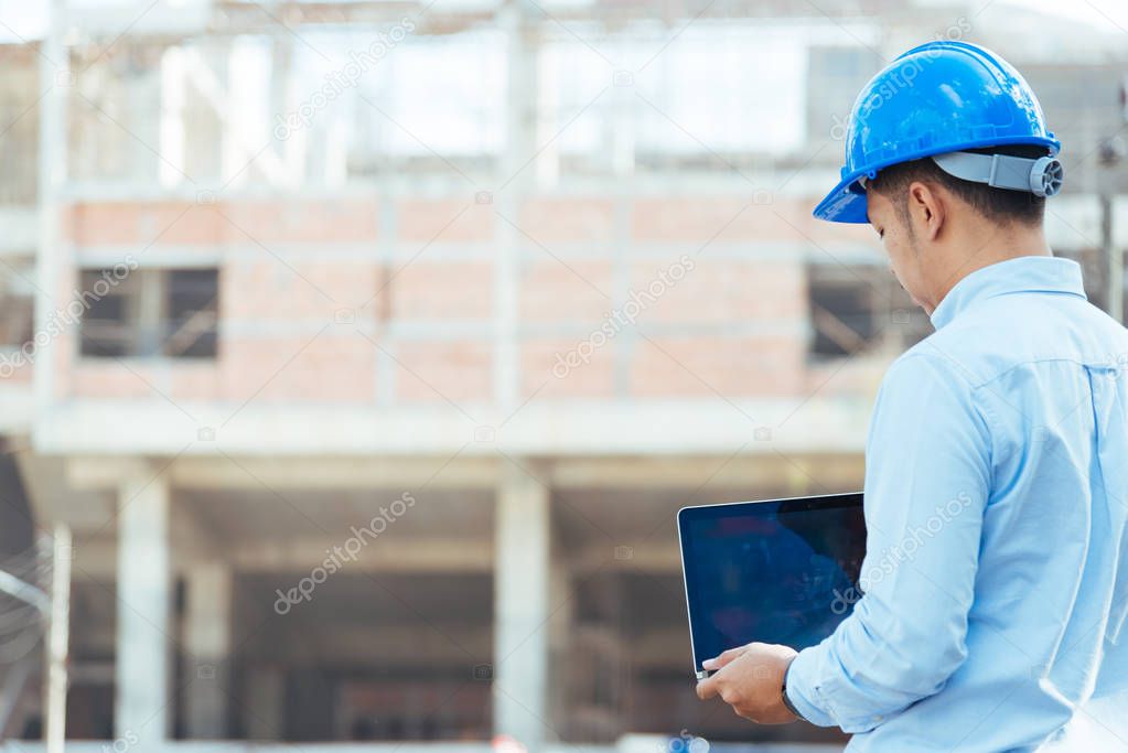 Engineer with blue safety helmet holding the laptop at the construction site.