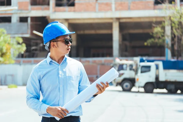 Civil engineer with blue safety helmet and sunglasses holding blueprint or drawing at construction site.