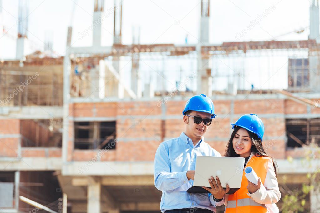 Asian man and woman engineer with the blue safety helmet meeting at the construction site with blueprints and laptop.