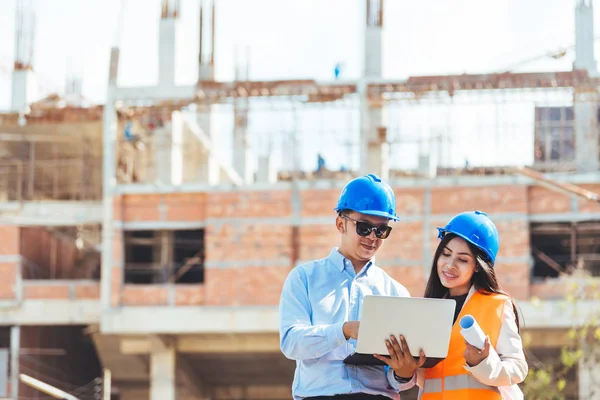 Asian man civil engineer holding laptop and Asian woman architect holding drawing blueprint at construction site.