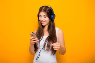 Çekici, akıllı telefonlu ve kulaklıklı genç bir kadın, sarı arka planda dururken müzik dinliyor..