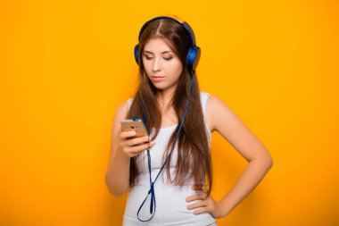 Çekici, akıllı telefonlu ve kulaklıklı genç bir kadın, sarı arka planda dururken müzik dinliyor..