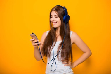 Çekici, akıllı telefonlu ve kulaklıklı genç bir kadın, sarı arka planda dururken müzik dinliyor..