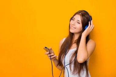 Çekici, akıllı telefonlu ve kulaklıklı genç bir kadın, sarı arka planda dururken müzik dinliyor..