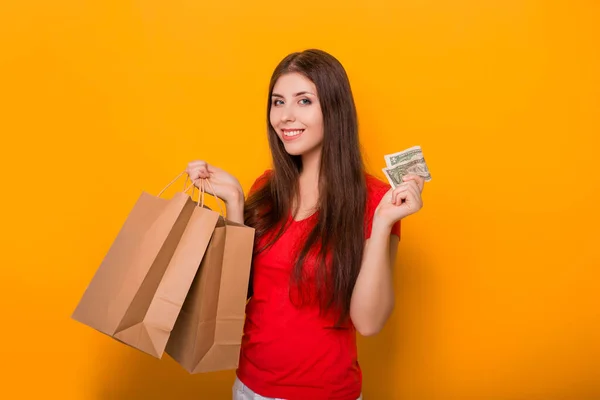 Attractive, young woman with money and packages in her hands, smiling while standing on a yellow background.