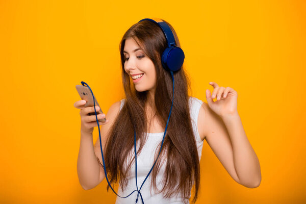 Attractive, young woman with a smartphone and in headphones, listens to music while standing on a yellow background.