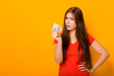 Genç bir kadının elinde 1 dolarlık banknot var ve kafası karışmış görünüyor. Küçük gelir kavramı. Sarı arka planda stüdyo fotoğrafı.