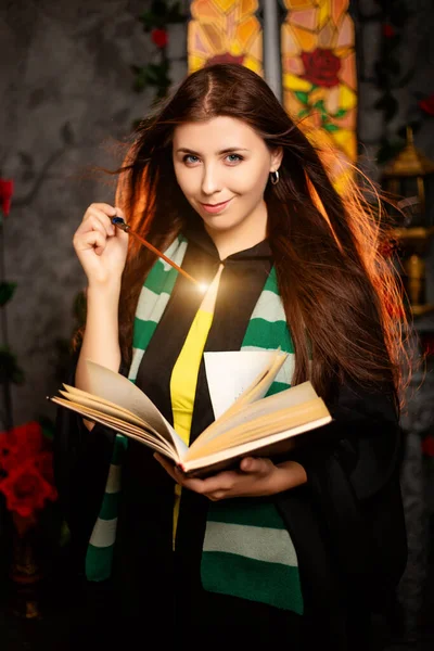 首に縞模様のスカーフをつけた黒いマントルの若い女性が本から呪文を唱え 魔法の杖で思いを馳せる — ストック写真