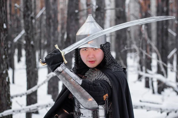 鎧を着た中世の戦士の肖像画 冬の森と雪を背景に彼の手にサーベルとヘルメットと黒のマント — ストック写真
