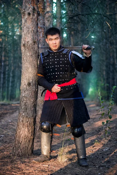 Warrior in black armor with a sword in his hands is leaning against a tree in a gloomy enchanted, damned forest.