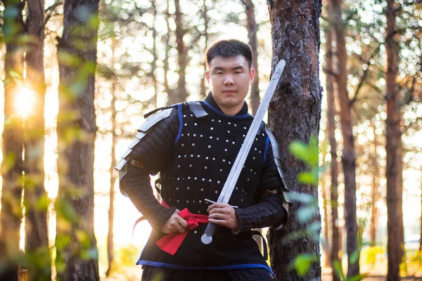 Warrior in black armor with a sword in his hands, is leaning against a tree in a gloomy forest.