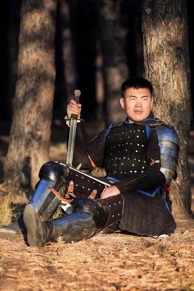 A warrior in black armor with a sword in his hands sits on the ground, leaning against a tree against the backdrop of a gloomy forest.
