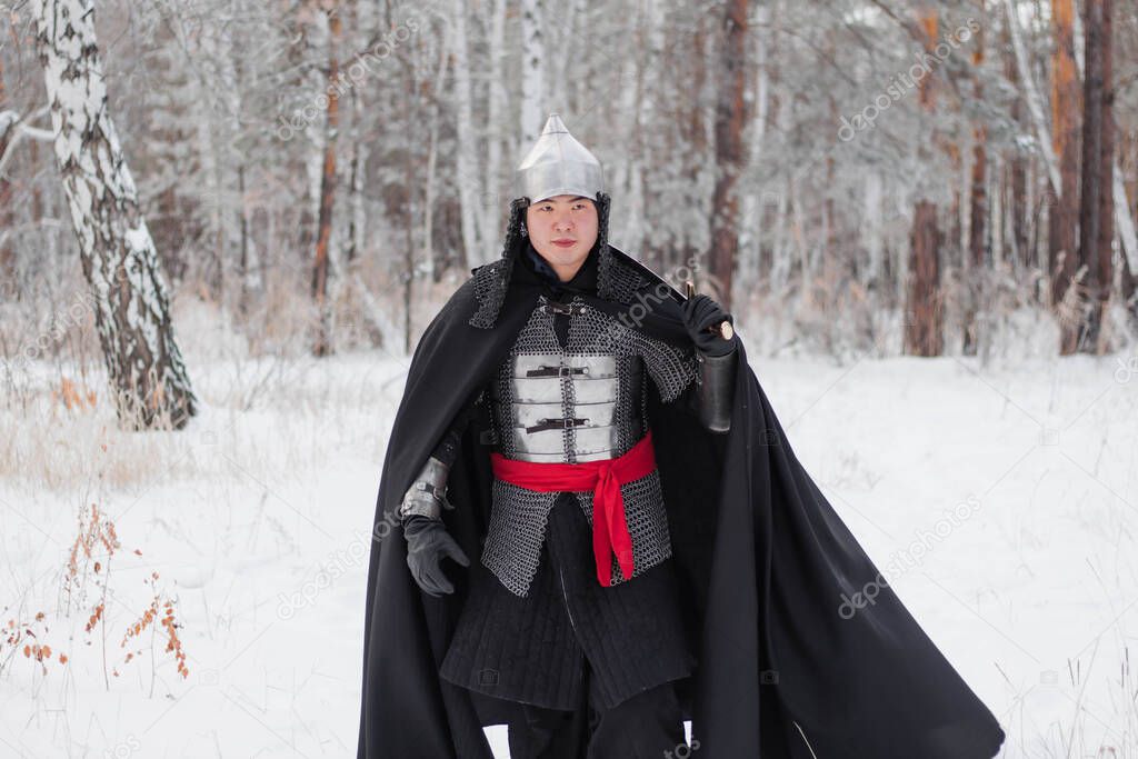 Medieval warrior in armor, helmet, black cloak with a saber in his hands walking in the winter in the forest.