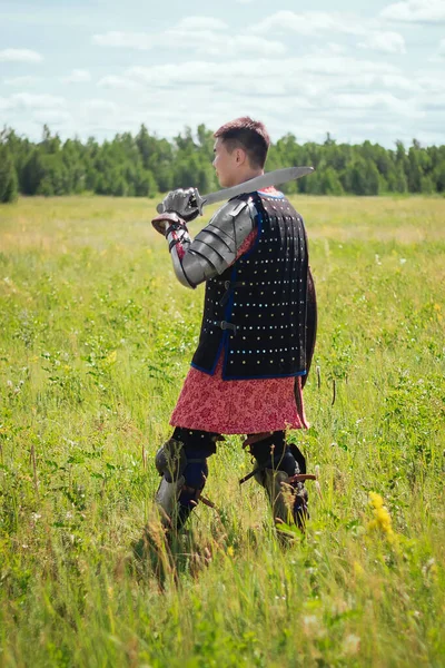 Steppe warrior in Mongolian armor of the 14th century in the field against the background of the forest and the blue sky. With a shield and a sword in his hands. Asian soldier nomad.