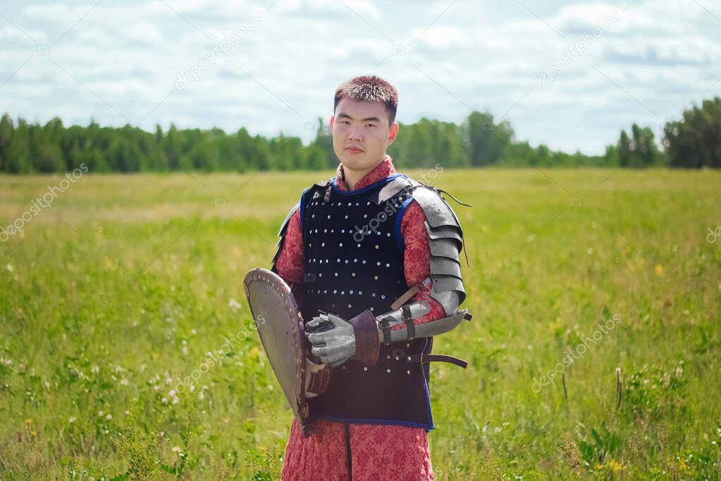 Steppe warrior in Mongolian armor of the 14th century in the field against the background of the forest and the blue sky. With a shield and a sword in his hands. Asian soldier nomad.