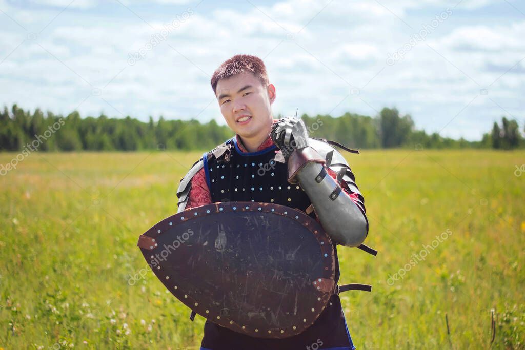 Steppe warrior in Mongolian armor of the 14th century in the field against the background of the forest and the blue sky. With a shield and a sword in his hands. Asian soldier nomad.