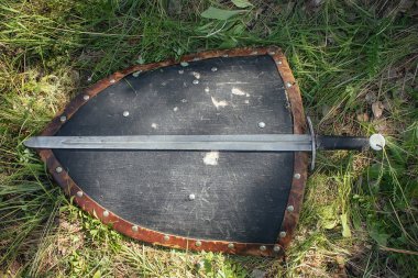 Kılıç üçgen bir kalkanda, yerde, uzun yeşil bir çimenlikte yatıyor. Savaştan sonra kaybettiğimiz silahlar, savaş alanı. Ortaçağ şövalyeleri..