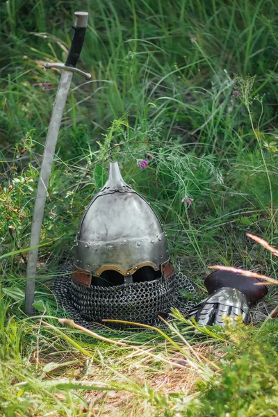 The helmet, sword and glove of a Mongolian warrior lie on the ground in a tall green grass. Lost, taken after the battle. Medieval weapons of a nomad, steppe soldier.