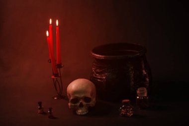 Kasvetli kırmızı ışık bir kafatasının üzerine düşüyor, bir kazan, mumlar ve şişeler karanlıkta siyah bir arka planda..