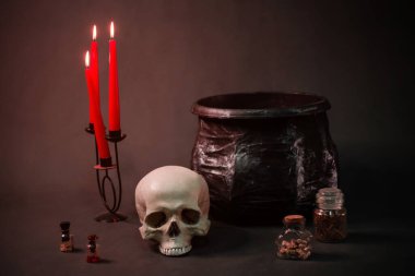 Kasvetli kırmızı ışık bir kafatasının üzerine düşüyor, bir kazan, mumlar ve şişeler karanlıkta siyah bir arka planda..