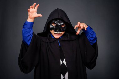 Siyah pelerinli, kapüşonlu ve siyah kafatası maskeli gri arka planda duran bir çocuk. Cadılar Bayramı kostümü giymiş bir çocuk tehditkar hareketler yapar, korkutur..