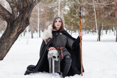  Kız, elinde mızrakla paltosu ve kürkü olan Viking döneminden bir savaşçı. Bir ağacın ve kış ormanının arka planında dizlerinin üzerinde oturuyor..